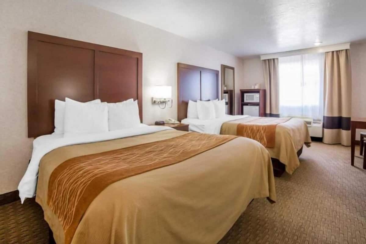 Une chambre d’hôtel avec deux lits, un bureau, une chaise, un miroir, une lampe et une fenêtre avec des rideaux.