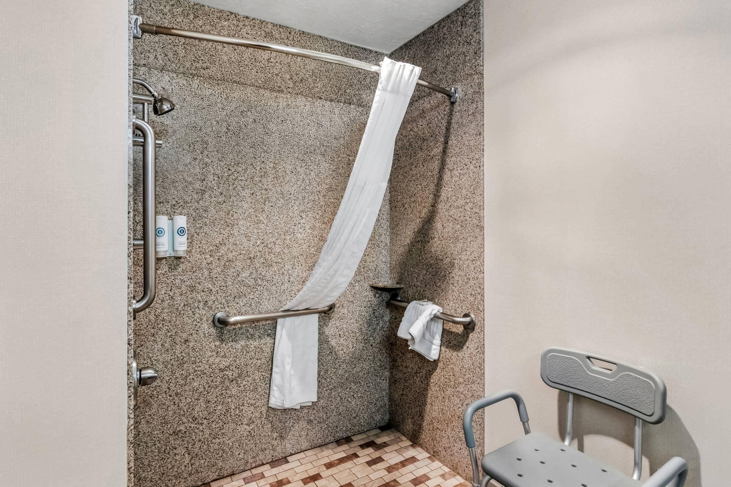 A bathroom with a shower, grab bars, a wheelchair-accessible chair, and a towel rack.