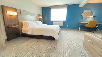 A modern hotel room with a bed, bedside table, desk, chair, and a large window.