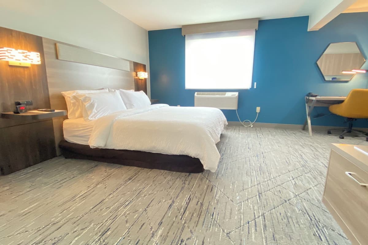 A modern hotel room with a bed, bedside table, desk, chair, and a large window.