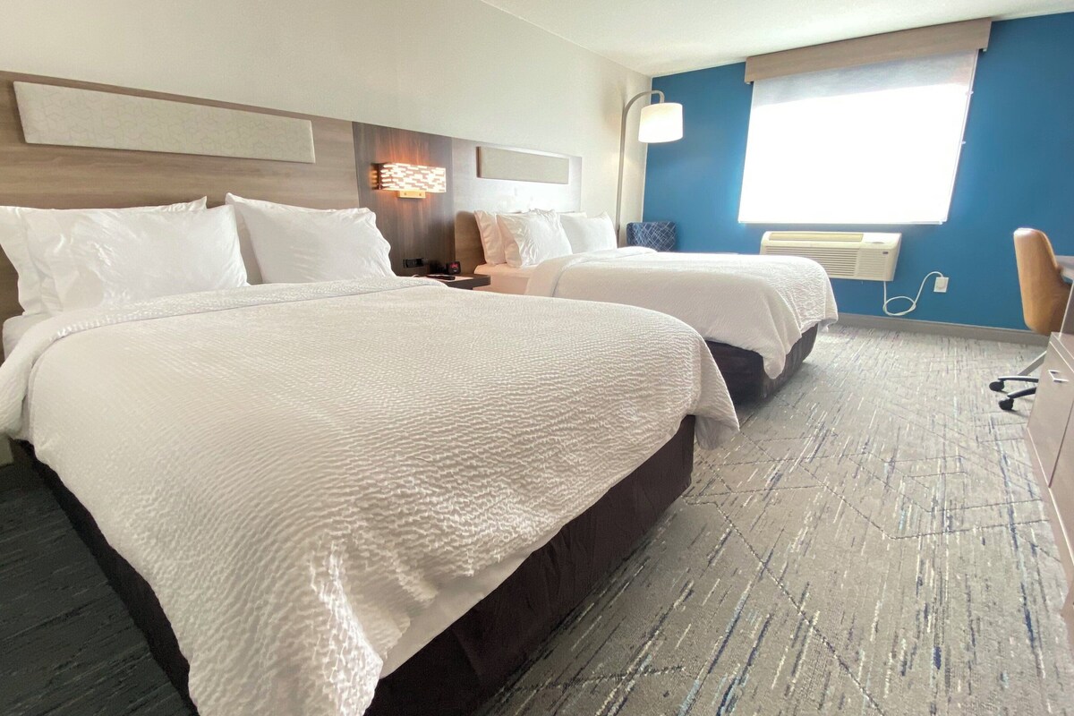 A hotel room with two beds, a desk, a chair, and a window with blinds.