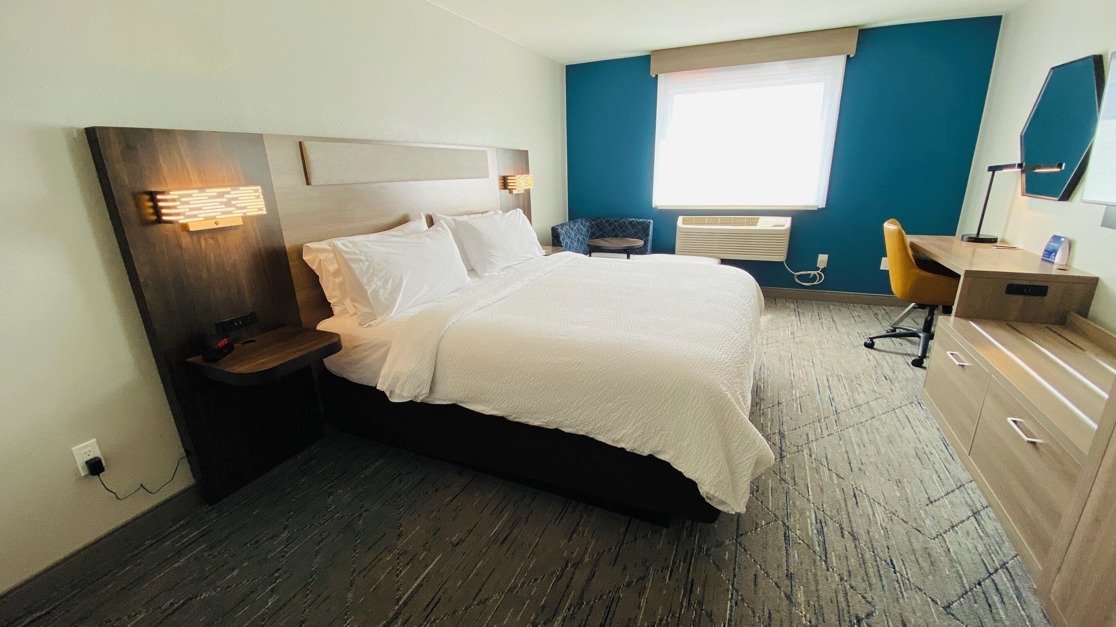 Habitación de hotel con cama, mesitas de noche, escritorio, silla, ventana con persianas y aire acondicionado instalado en la pared.