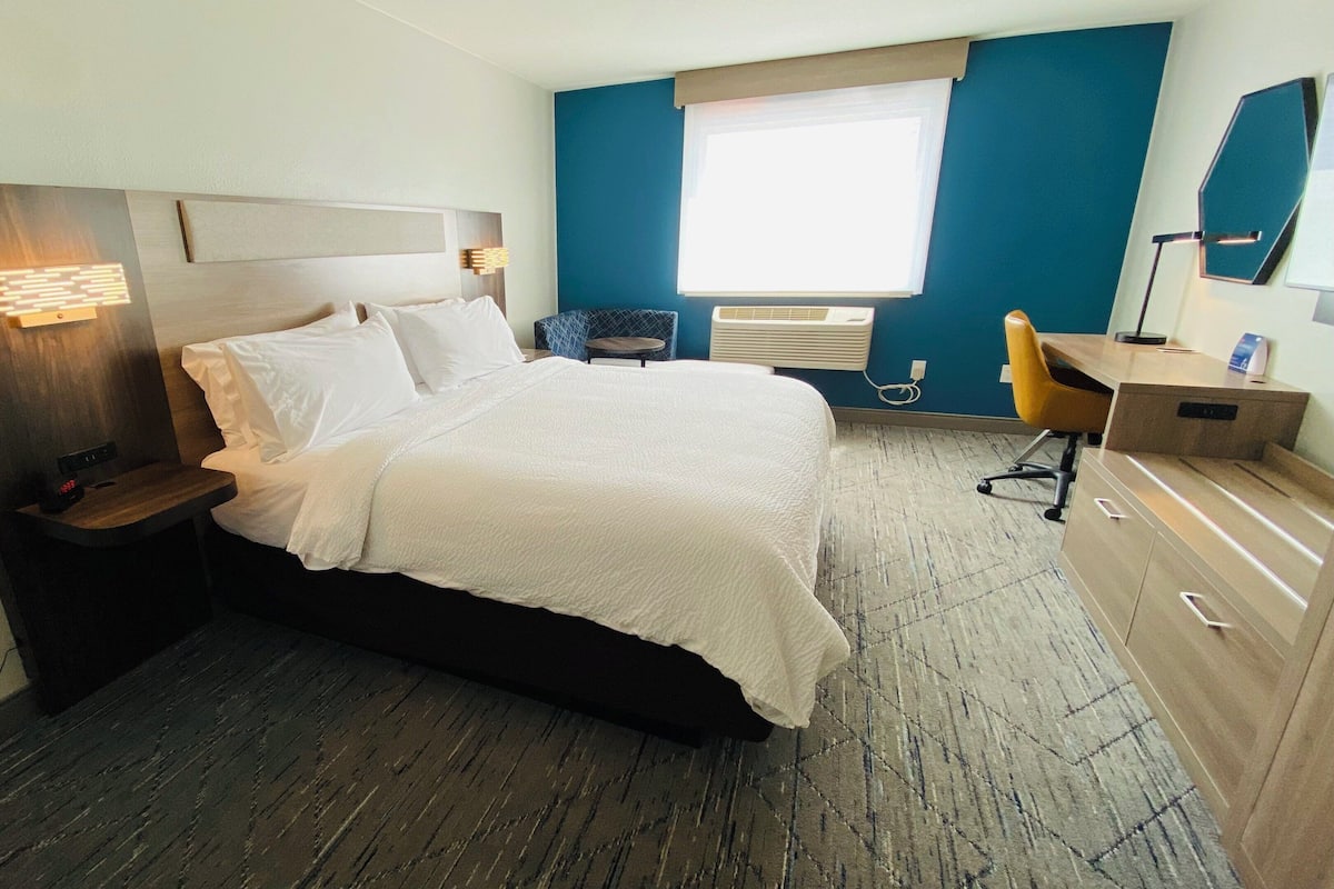 A hotel room with a bed, bedside tables, a desk, a chair, a window with blinds, and a wall-mounted air conditioning unit.