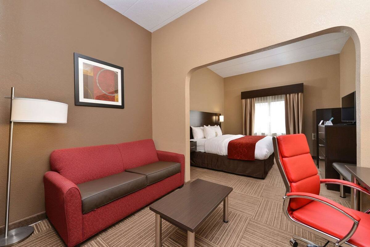A hotel room with a red sofa, a small table, a red chair, a bed with white linens, a TV, and a framed picture on the wall.
