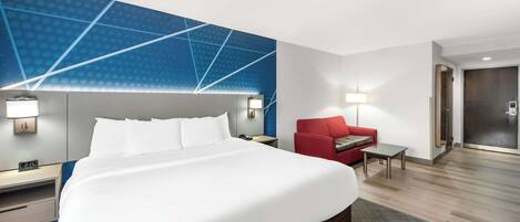 Habitación de hotel con una cama grande, un sillón rojo, una mesita pequeña y una pared con un diseño geométrico.