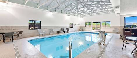 An indoor swimming pool with clear blue water, surrounded by lounge chairs and a high ceiling with metal beams.