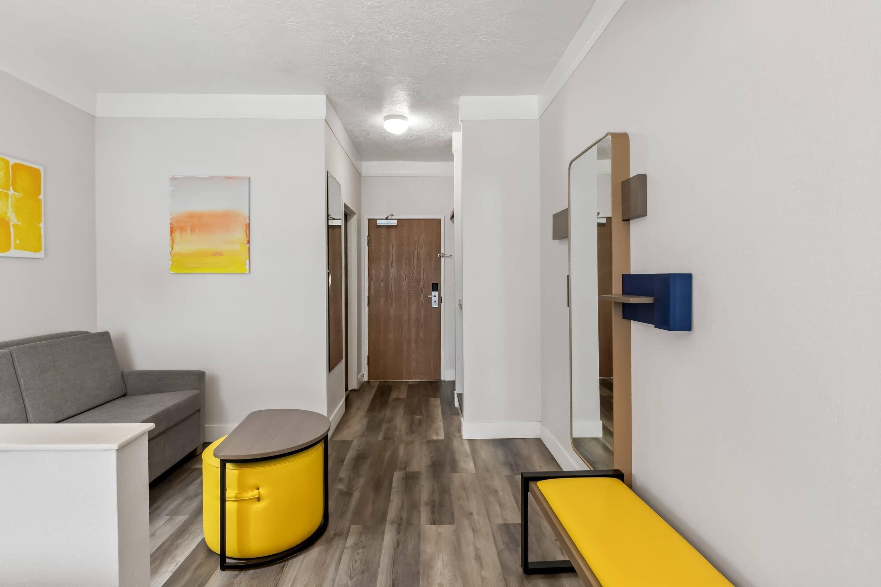 Une chambre d’hôtel moderne avec un canapé gris, un pouf jaune, une petite table et un banc.