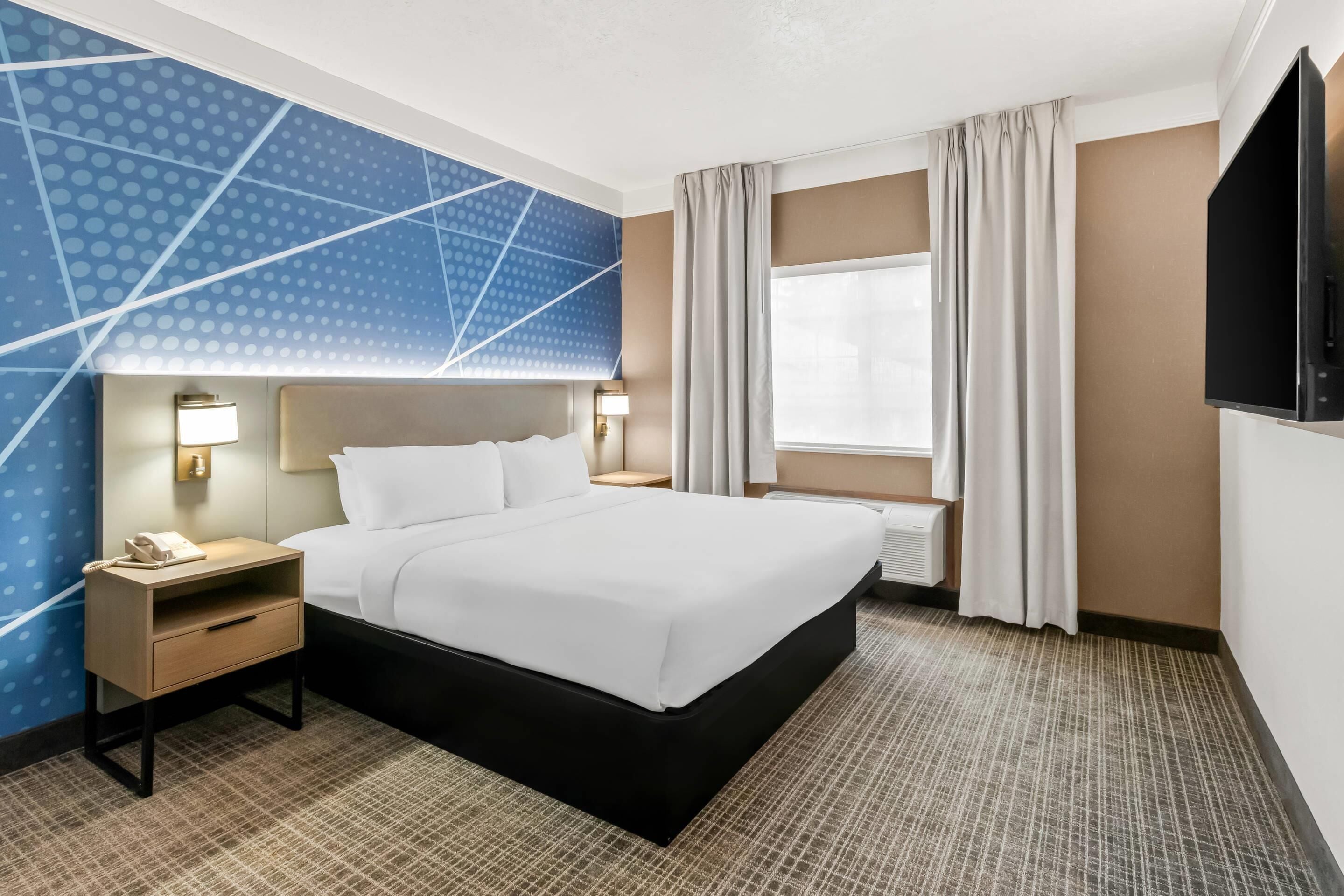 Una habitación de hotel con cama, mesitas de noche, televisor y ventana con cortinas.