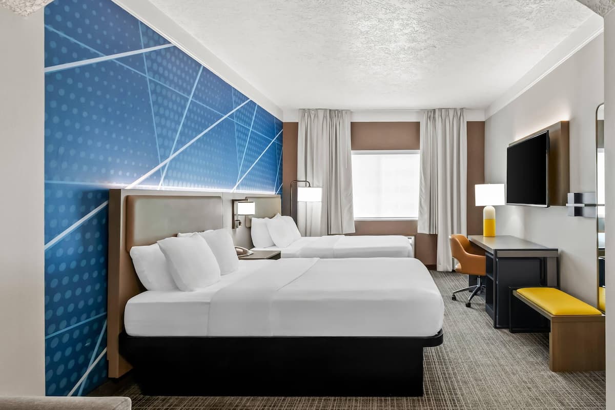 A hotel room with two beds, a desk, a chair, a TV, and a large window with curtains.