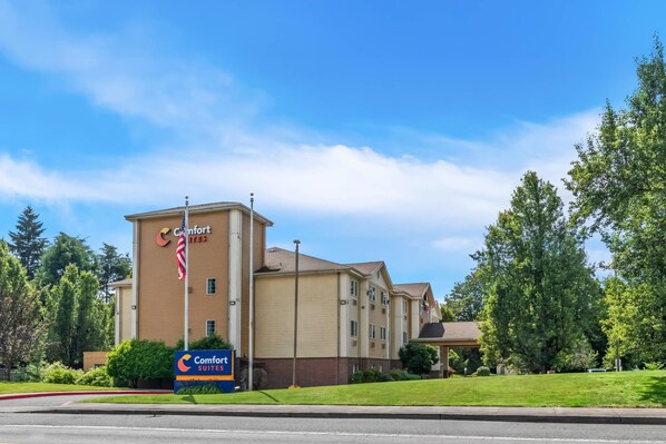 A Comfort Suites hotel with a beige exterior, a flagpole, and a signboard. - Comfort Suites Clackamas (Clackamas)