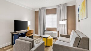 A modern hotel room with a flat-screen TV, a desk, a yellow ottoman, and a sofa set. - Comfort Suites Clackamas (Clackamas)