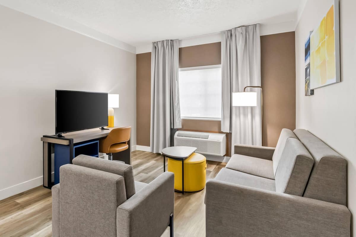 A modern hotel room with a flat-screen TV, a desk, a yellow ottoman, and a sofa set.