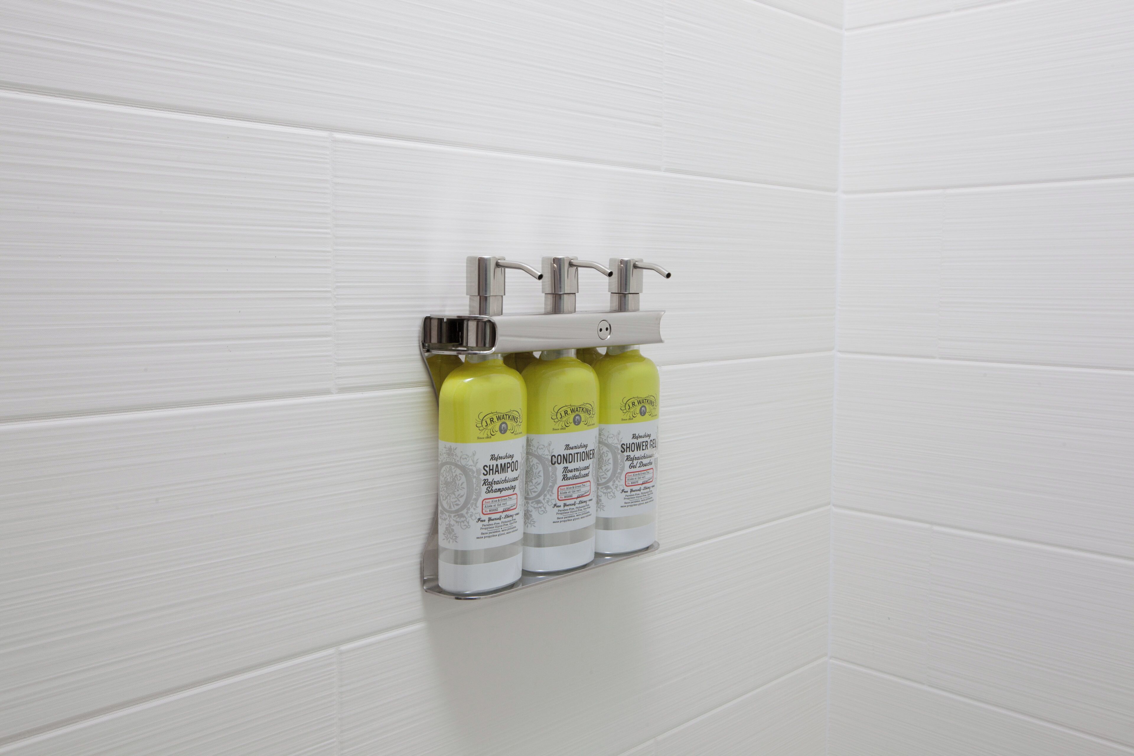A bathroom with white tiled walls, a chrome faucet, and three bottles of J.R. Watkins shower gel.