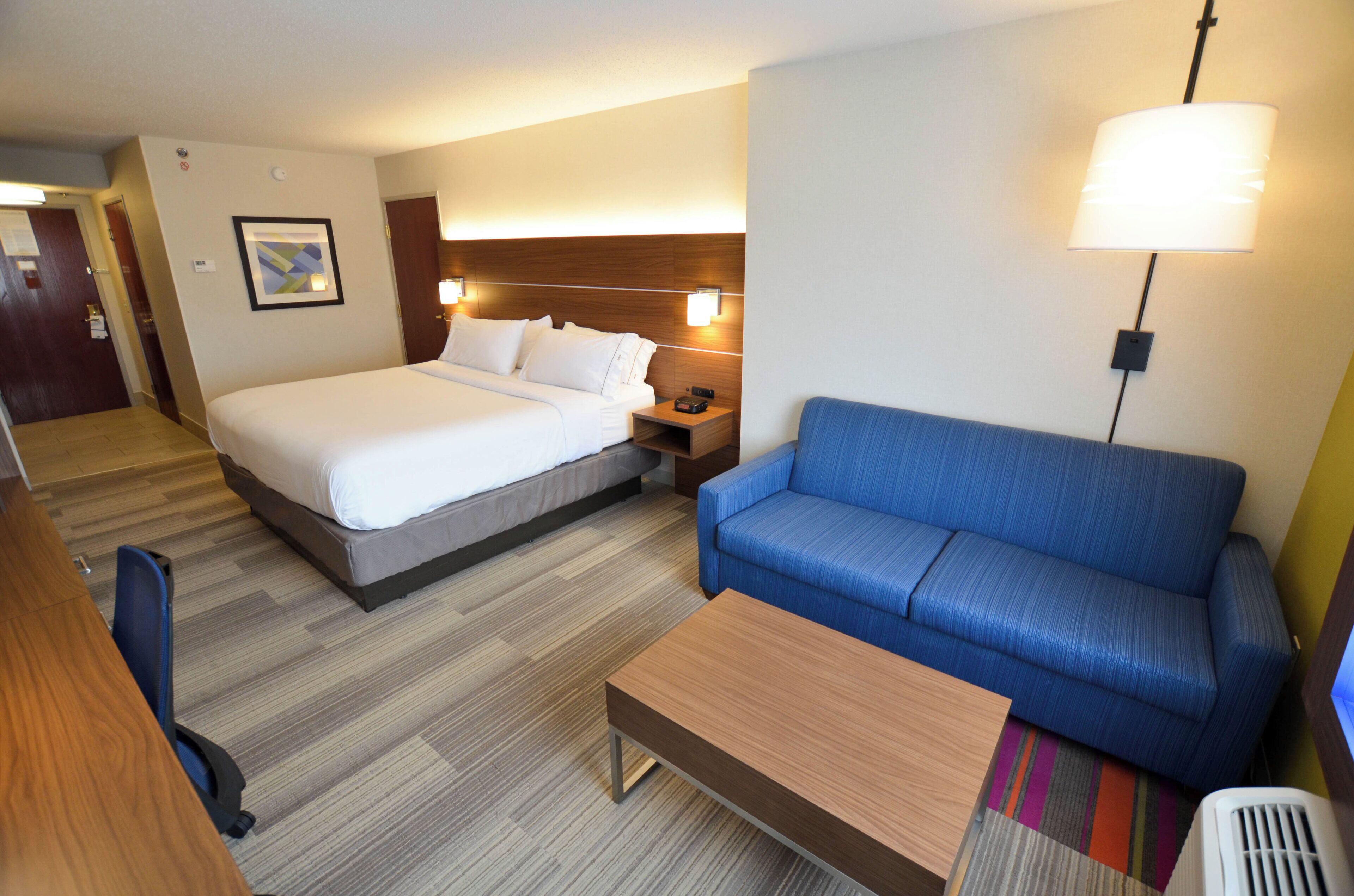 A hotel room with a bed, a blue sofa, a wooden coffee table, a desk with a chair, and a wall-mounted lamp.