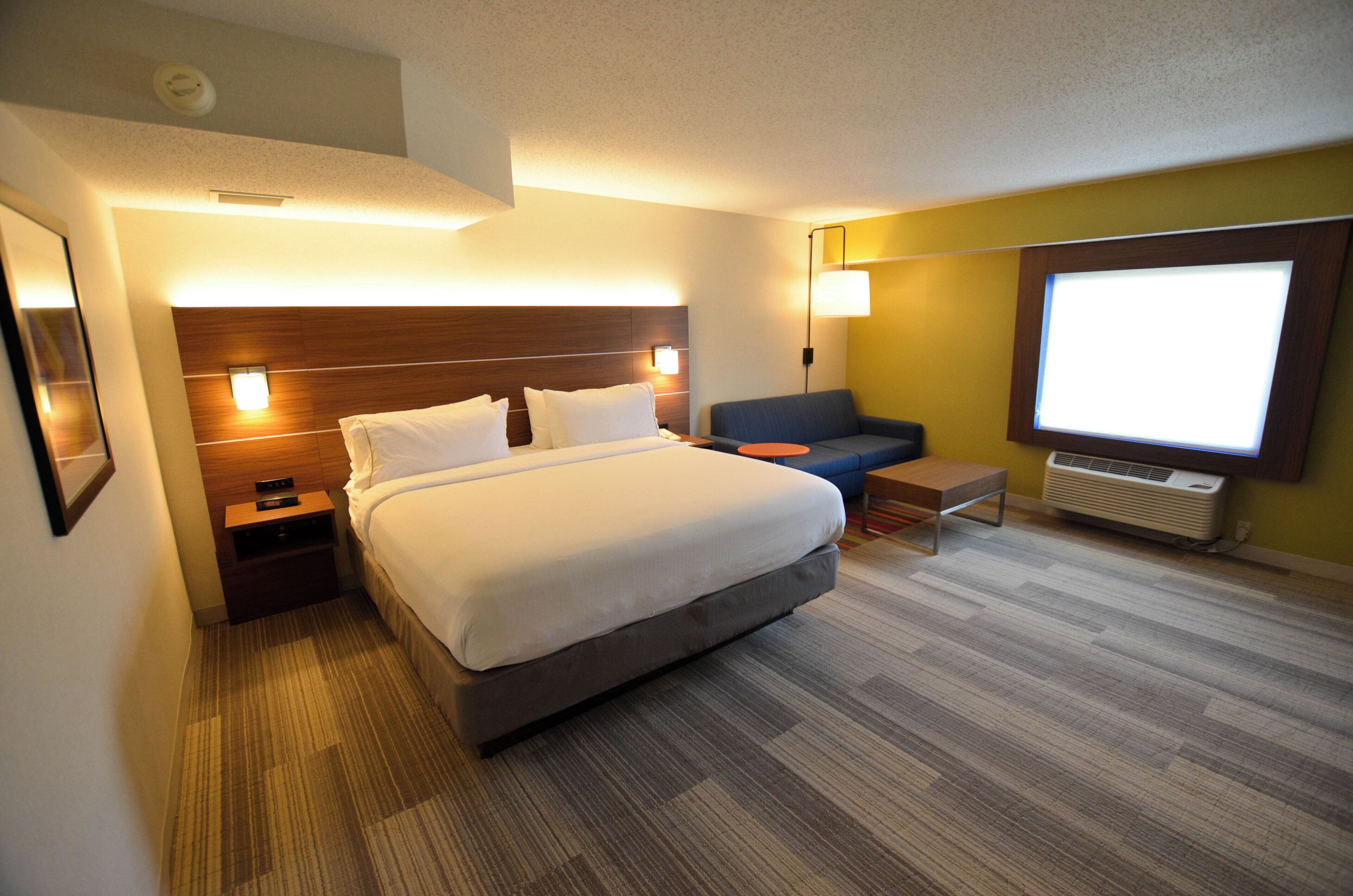 A hotel room with a bed, a desk, a chair, a TV, and a wall-mounted heater.