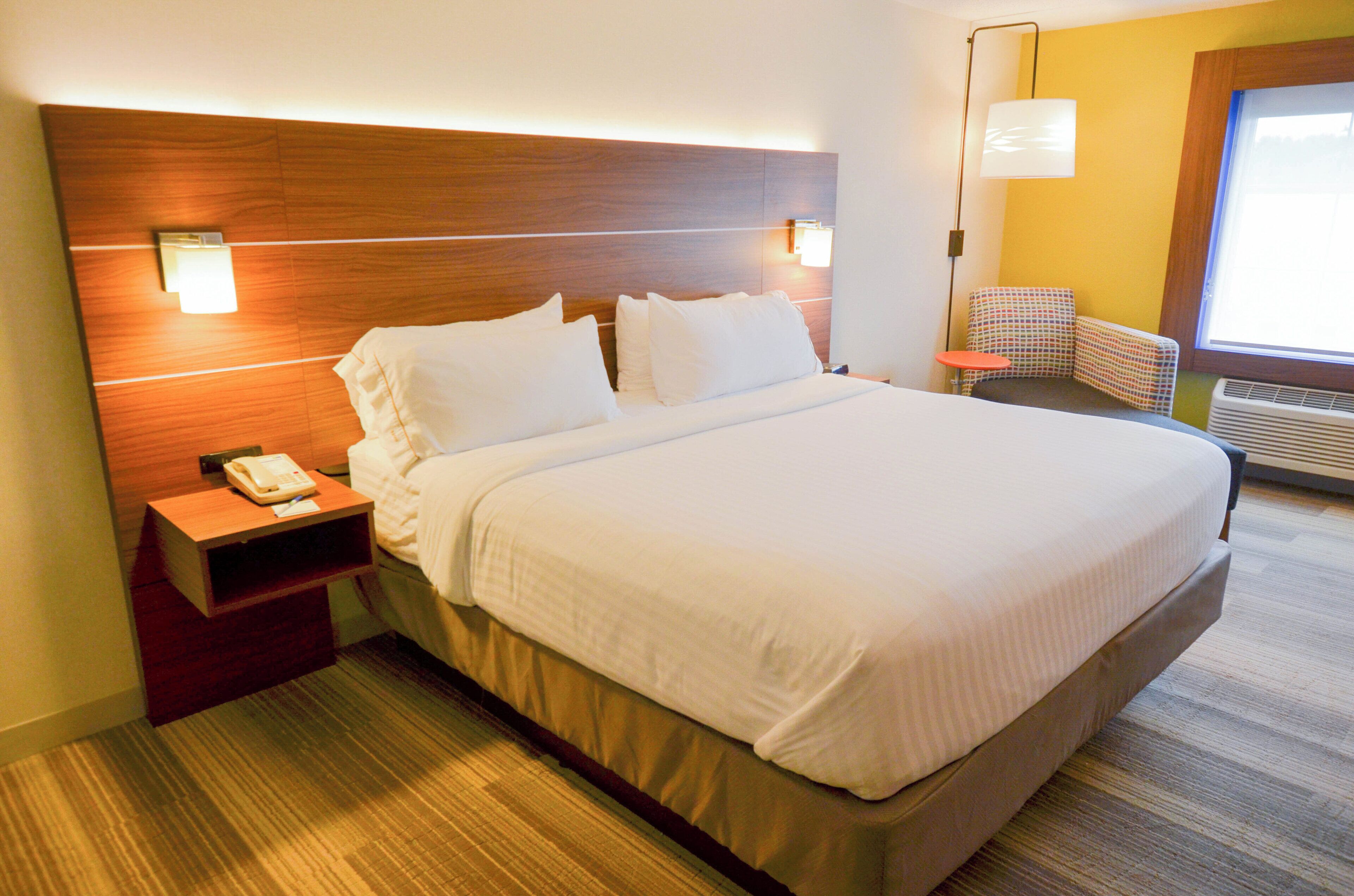 A hotel room with a large bed, a bedside table with a telephone, a small table with a chair, and a window with blinds.