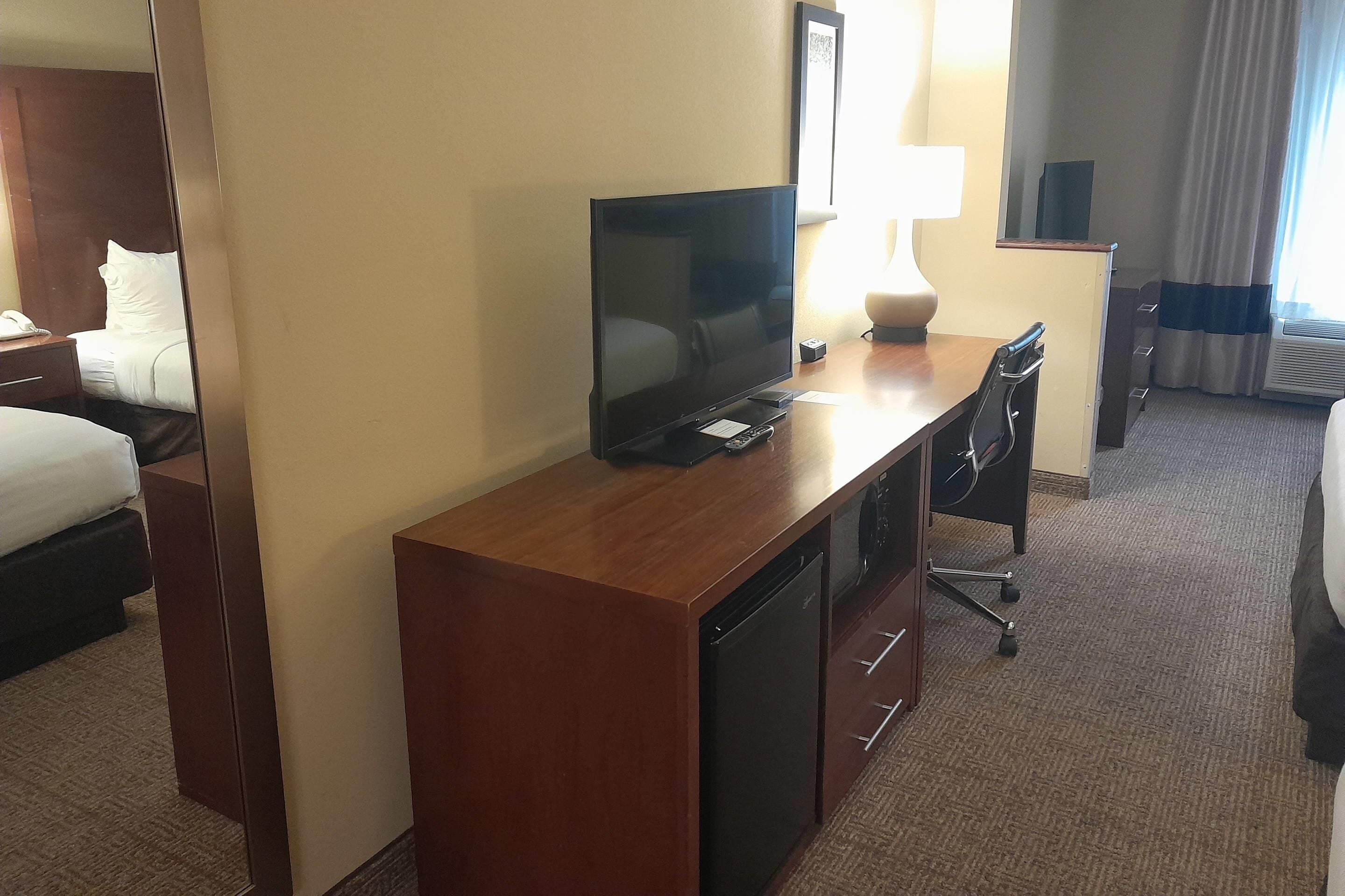 A hotel room with a wooden desk, a flat-screen TV, a lamp, a mini-fridge, a desk chair, and a bed with white bedding.