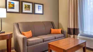 A hotel room with a grey sofa, two orange cushions, a wooden coffee table, and three framed pictures on the wall. - Comfort Inn Duncansville - Altoona (Hollidaysburg)