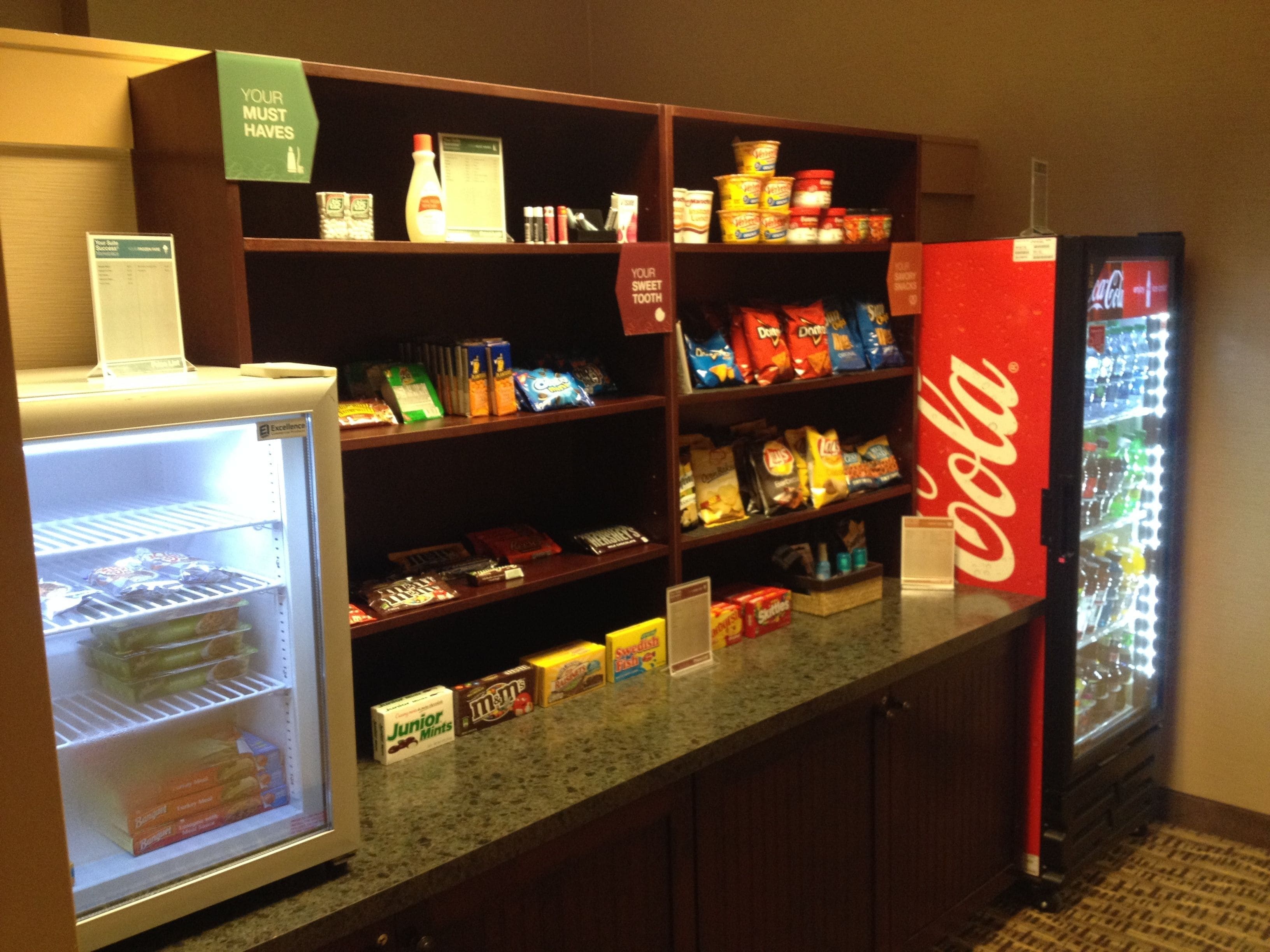 A self-service snack and beverage station with a refrigerator, shelves stocked with snacks, and a Coca-Cola refrigerator.