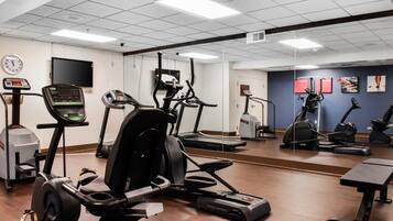 Et fitnessrum med forskellige konditionsmaskiner, en vægtbænk og et digitalt ur på væggen.