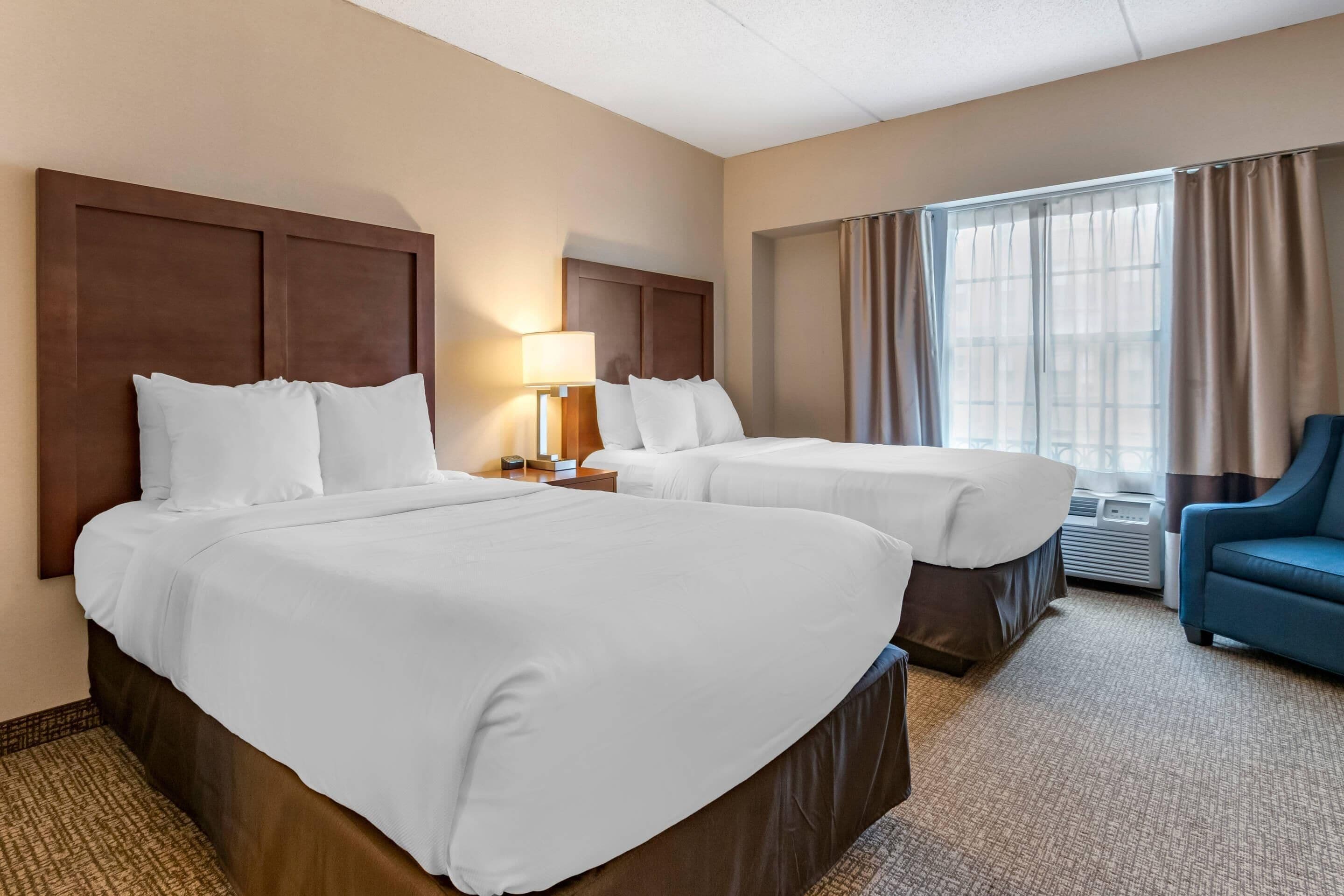 Een hotelkamer met twee bedden, een bureau met lamp, een stoel en een groot raam met gordijnen.