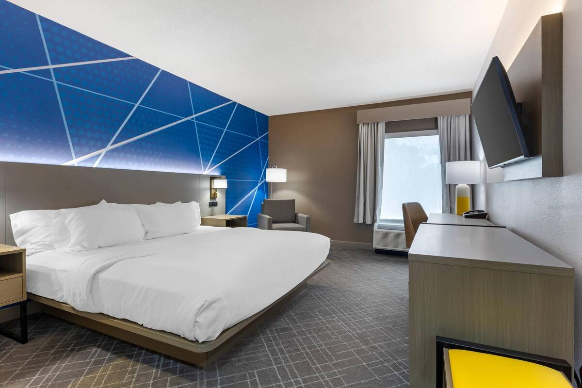 Habitación de hotel con una cama grande, un escritorio con silla, un televisor montado en la pared y una ventana con cortinas.