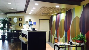 Uma receção com um computador, um relógio e um aviso com horários de check-in. Uma parede colorida com designs abstratos e uma planta num vaso sobre uma mesa.