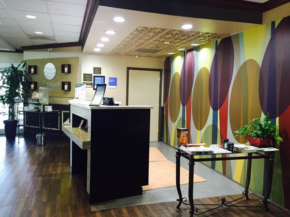 A reception desk with a computer, a clock, and a sign indicating check-in times. There is a colorful wall with abstract designs and a potted plant on a table.