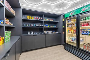 Un magasin bien organisé, avec des étagères remplies de divers produits, un rayon réfrigéré pour les boissons et un récipient en plastique transparent posé sur le comptoir.