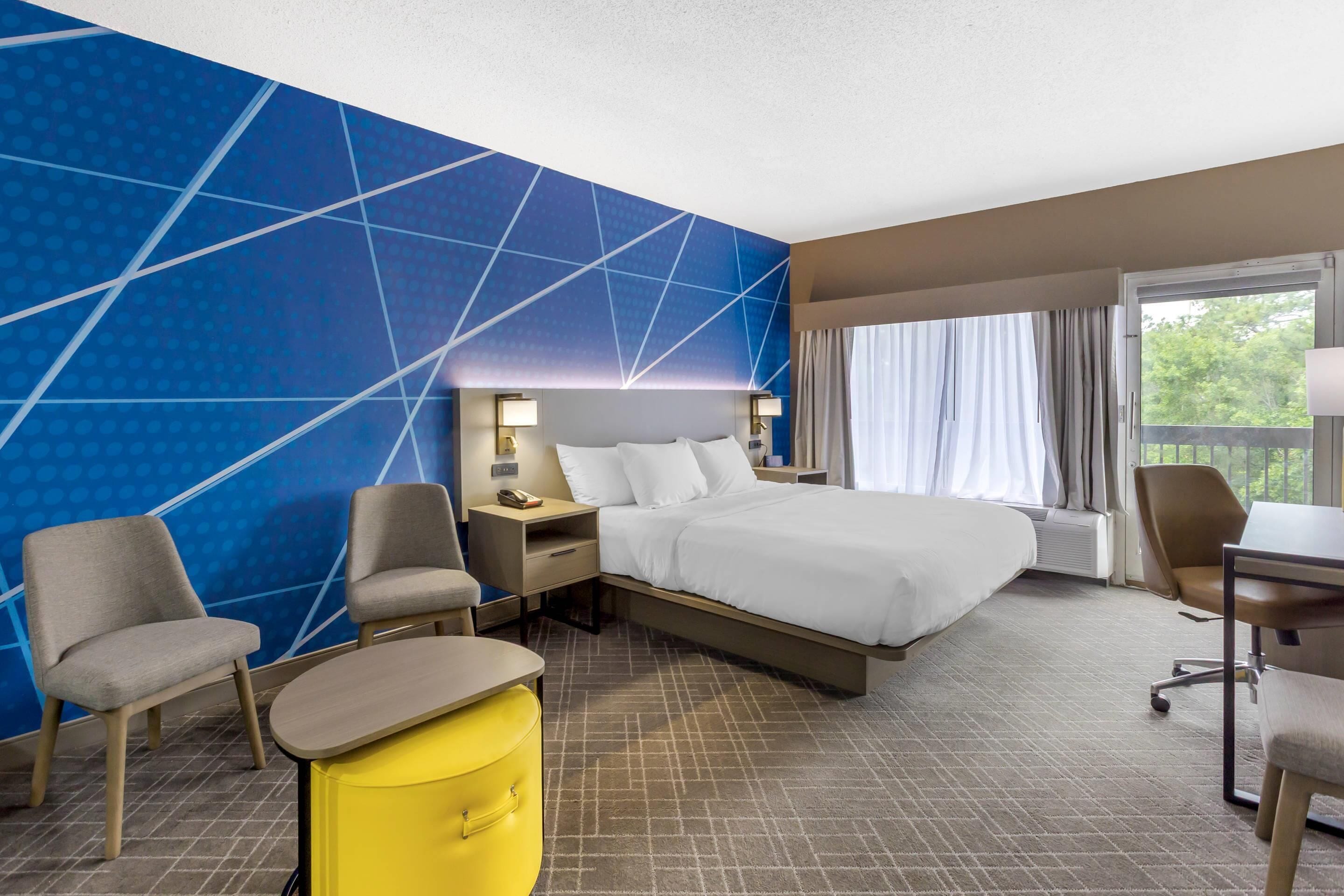 A hotel room with a large bed, two bedside tables with lamps, a desk, two chairs, and a yellow ottoman.