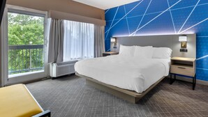Quarto de hotel com uma cama grande, mesa de cabeceira, cortinas e vista para árvores.
