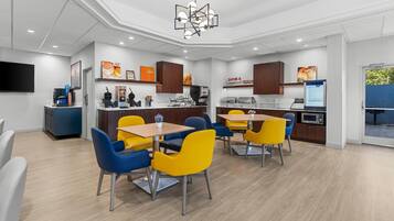 Un comedor moderno con una mesa central y sillas, una zona de cocina con electrodomésticos y un televisor de pantalla plana montado en la pared.