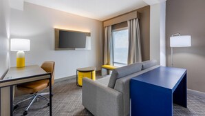Quarto de hotel com escrivaninha, cadeira, sofá e televisão fixada na parede.