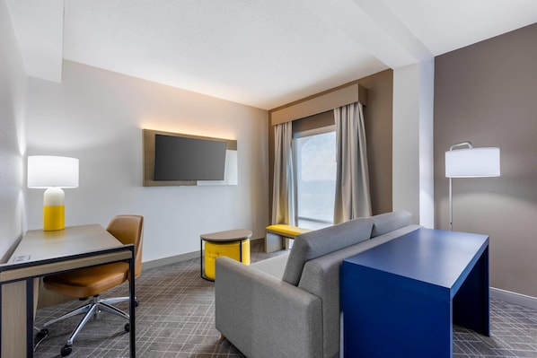 A hotel room with a desk, chair, sofa, and a TV mounted on the wall.