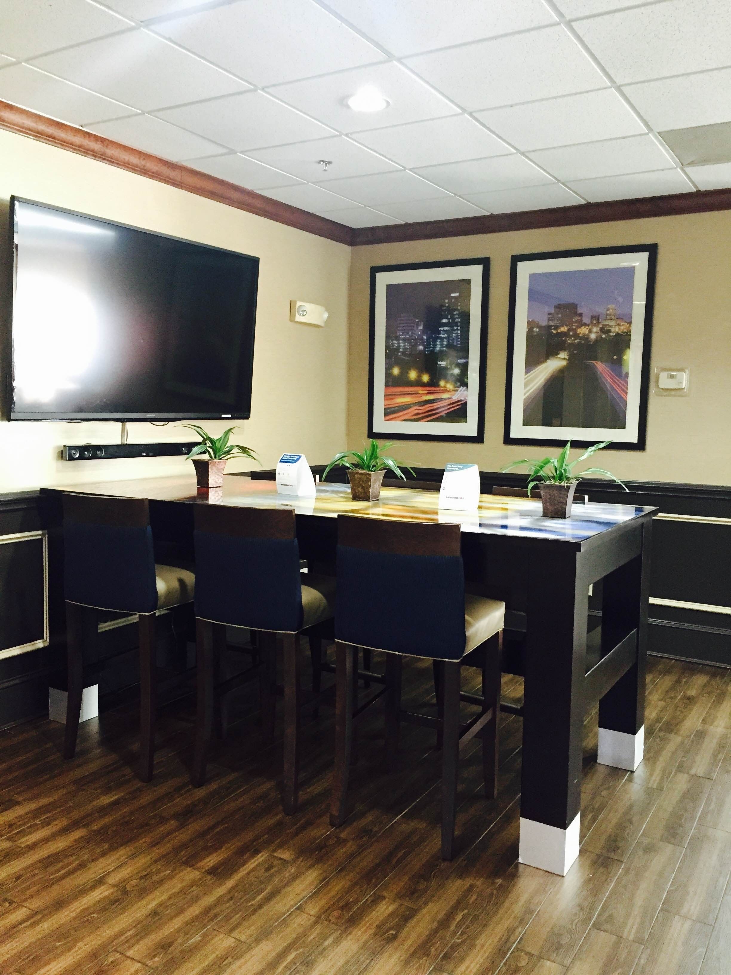 A conference room with a large flat-screen TV, a wooden table, and four chairs. There are two framed pictures on the wall and potted plants on the table.