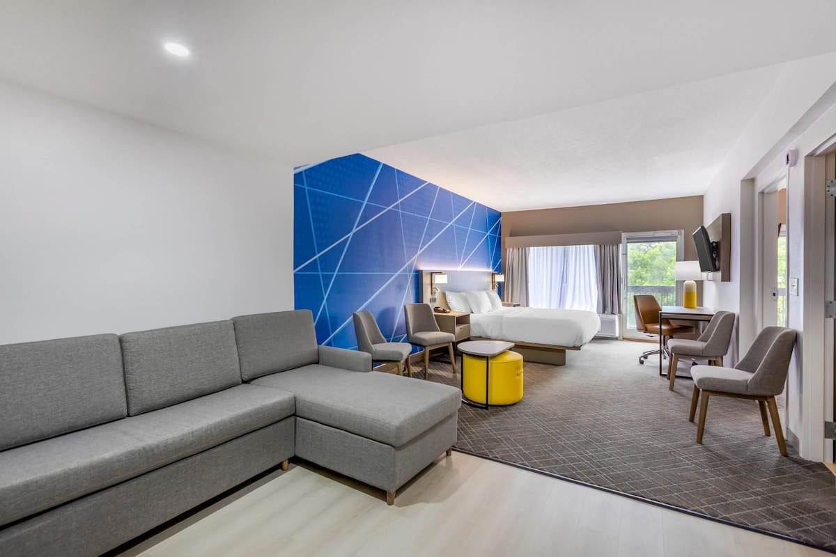 Una habitación de hotel moderna con un sofá seccional gris, un otoman amarillo redondo, un escritorio con dos sillas, una cama con ropa de cama blanca y un ventanal con cortinas.