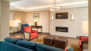 Un vestíbulo moderno de hotel con un sofá azul, sillones rojos, una chimenea y un televisor de pantalla plana montado en la pared.