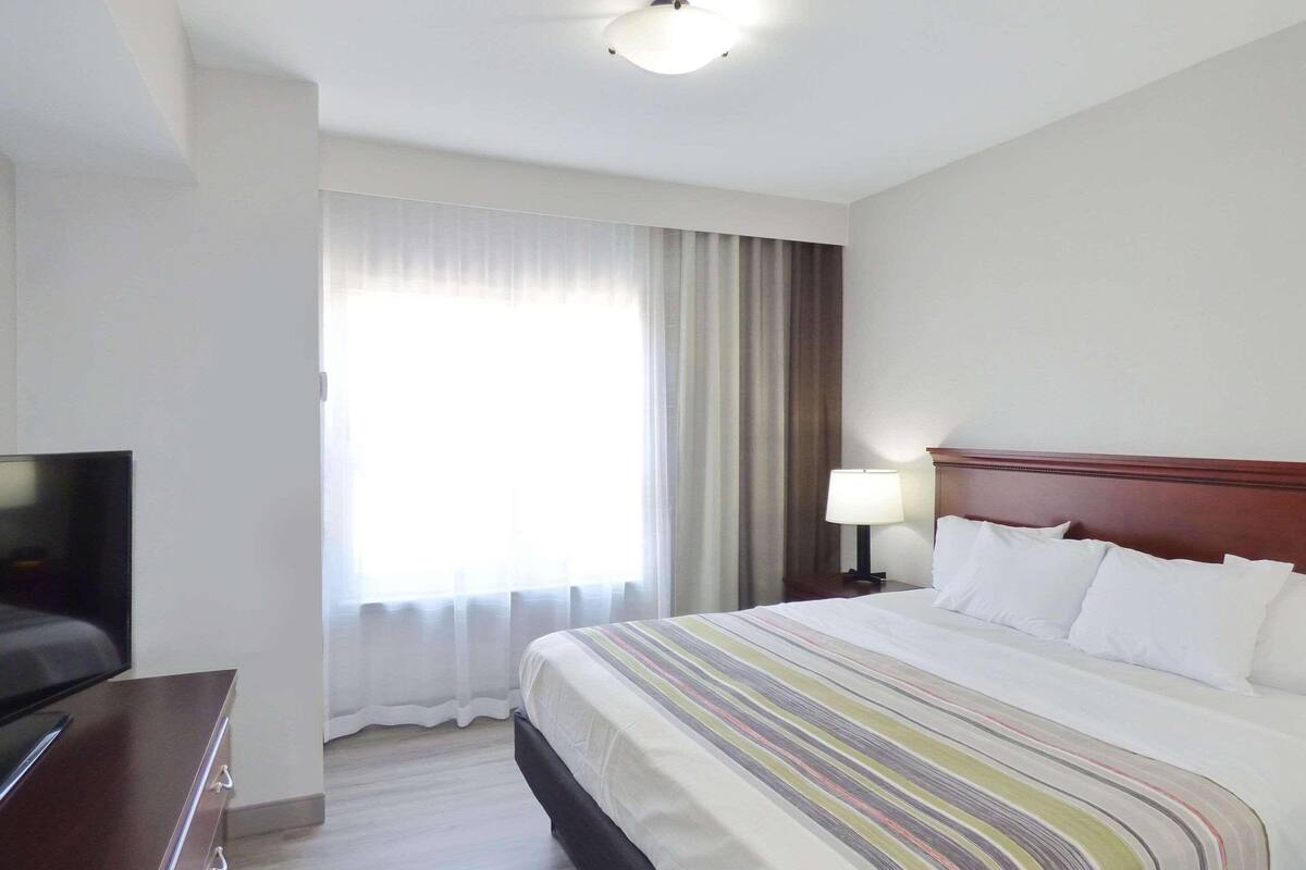 A hotel room with a bed, a TV, a nightstand with a lamp, and a window with curtains.