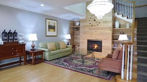 A living room with a fireplace, wooden floors, a green sofa, a coffee table, and a staircase with a chandelier. - Country Inn & Suites by Radisson, Biloxi-Ocean Springs, MS (Ocean Springs)