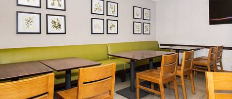 A restaurant interior with wooden tables and chairs, a green cushioned bench, and framed botanical artwork on the walls.
