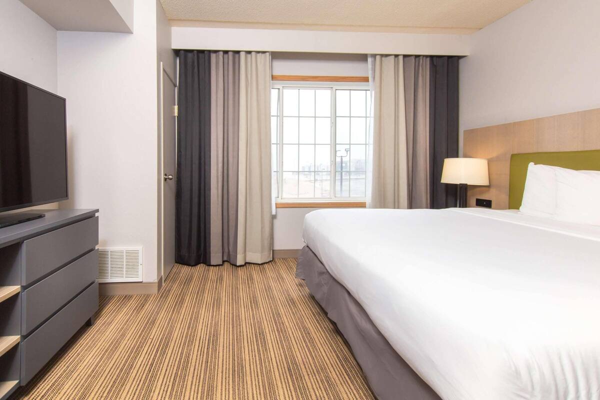 A hotel room with a bed, a television, a dresser, a window with curtains, and a lamp.