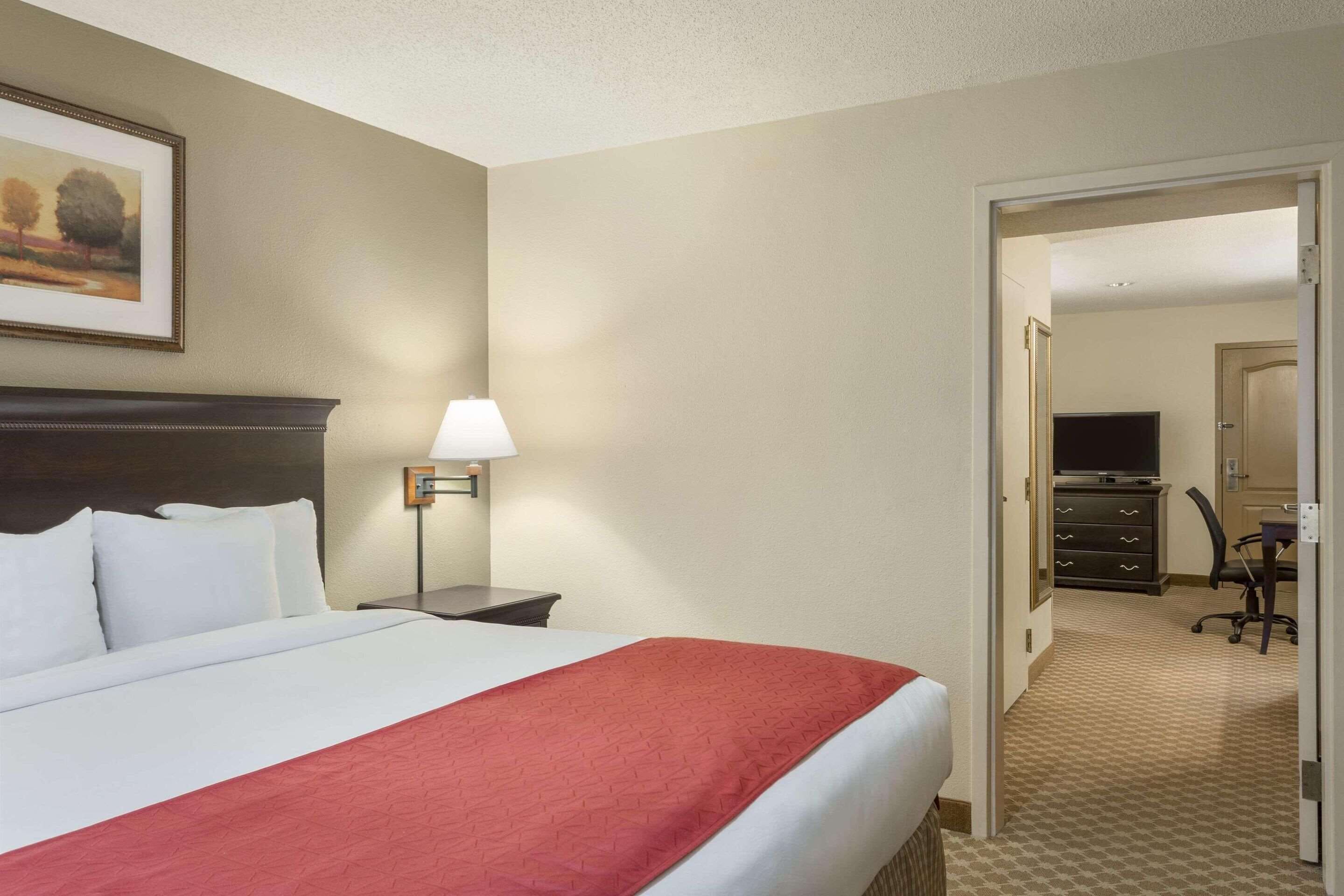 A hotel room with a large bed, a nightstand with a lamp, a dresser, and a television in the adjacent room.
