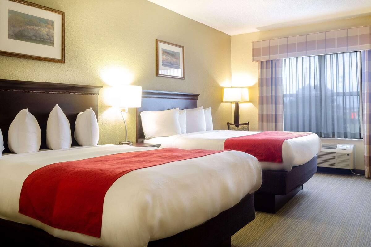 A hotel room with two beds, each with white linens and red throw blankets. There are two bedside lamps, a framed picture on the wall, and a window with curtains.