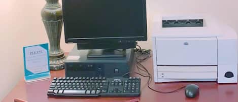 A desk with a computer, keyboard, mouse, and printer. There is a chair with a green seat cushion, a lamp, and a framed picture on the wall.