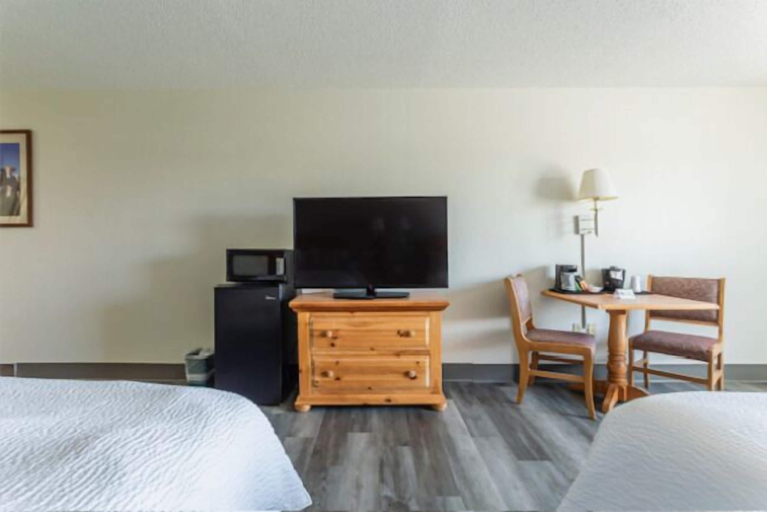 Quarto de hotel com duas camas, uma televisão, uma cômoda de madeira, uma escrivaninha com abajur e uma cadeira.