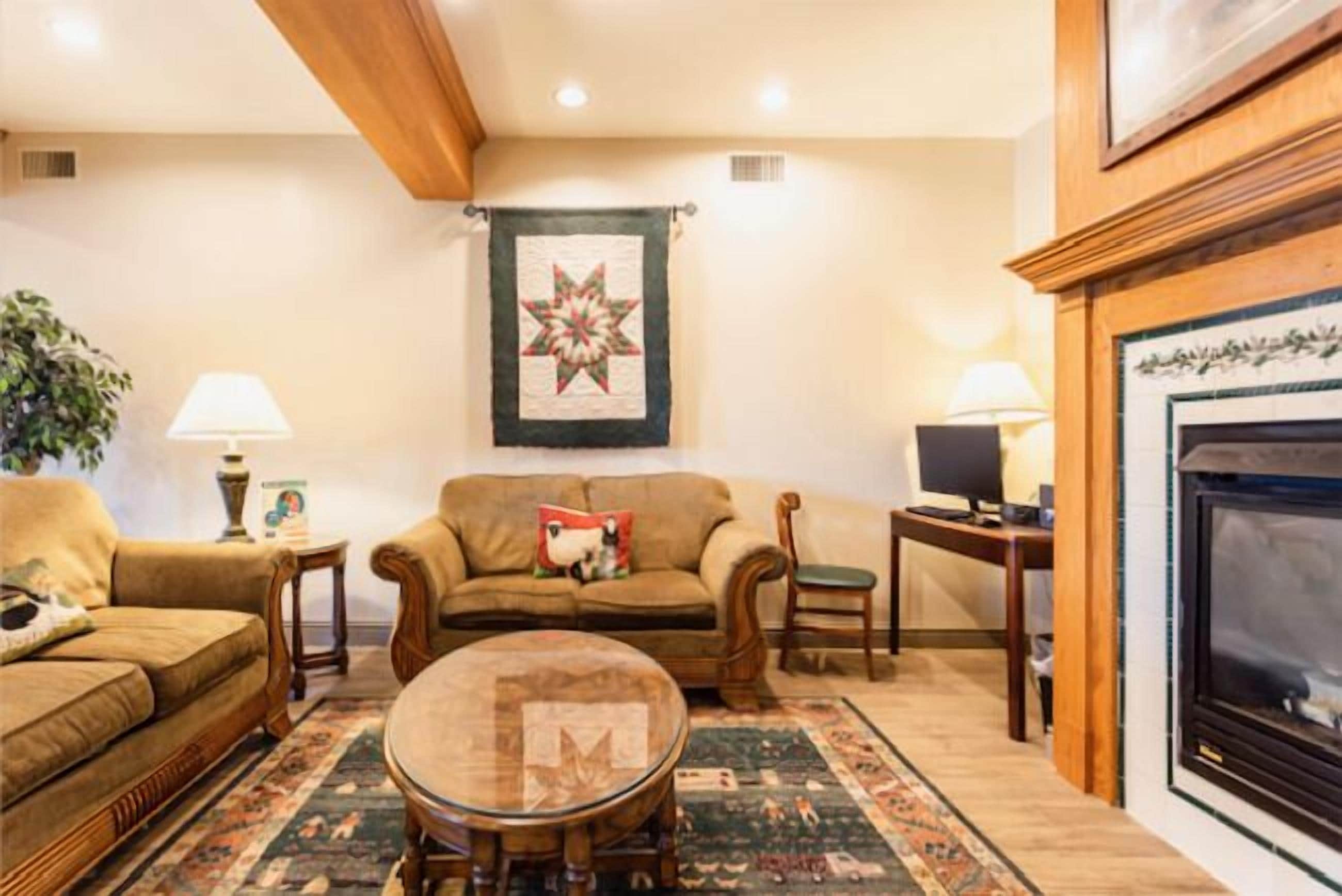 A cozy living room with a fireplace, a large quilt hanging on the wall, and a round coffee table.