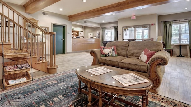 A spacious living room with a wooden staircase, a sofa with decorative pillows, and a round coffee table with newspapers.