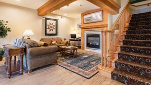A living room with a fireplace, a staircase with patterned carpet, and a wooden ceiling beam.