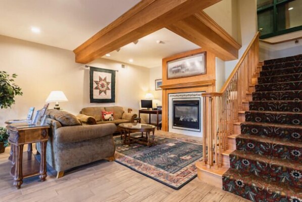 A living room with a fireplace, a staircase with patterned carpet, and a wooden ceiling beam.
