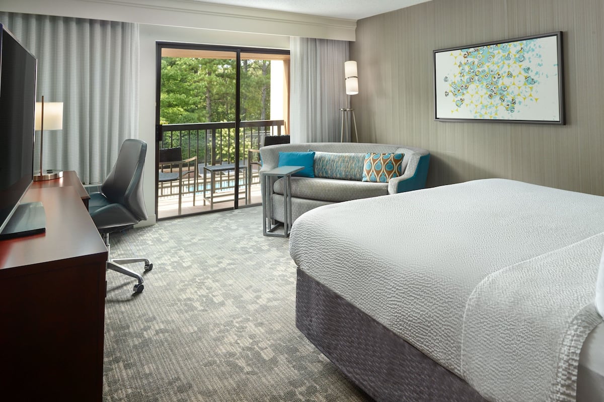 A hotel room with a large bed, a desk with a chair, a sofa, and a balcony with a view of trees.